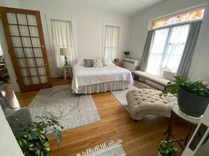 A bedroom with a bed, bedside tables, a window with curtains, a radiator, a potted plant, and a wooden floor.
