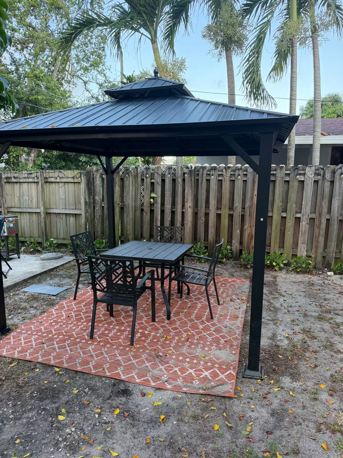 Un patio cubierto con una mesa y sillas, una cerca de madera al fondo y un cielo despejado.