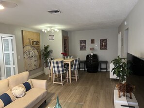 Ein Wohnzimmer mit einem beige farbenen Sofa, einem Esstisch mit Stühlen, einem Fernseher und einer Wanduhr.