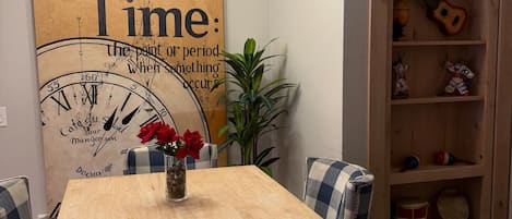Un comedor con una mesa de madera y sillas a cuadros, un reloj de pared y una estantería con diversos objetos.