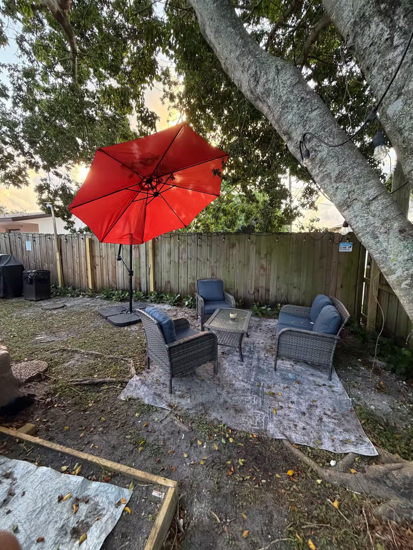 A backyard with a red umbrella, wicker furniture, and a wooden fence.