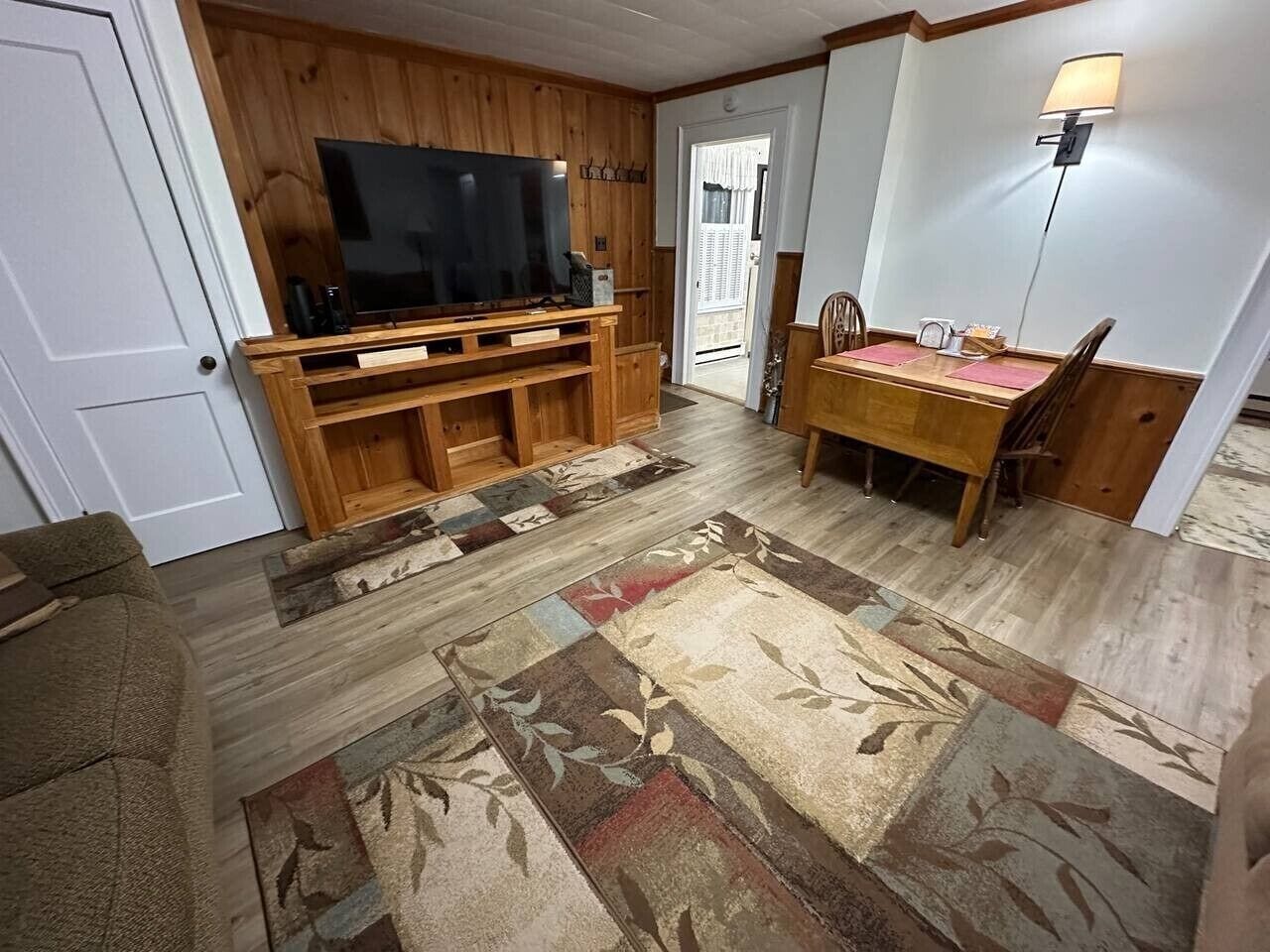 Un salon avec un téléviseur posé sur un meuble en bois, une table à manger avec des chaises et un tapis à motifs.
