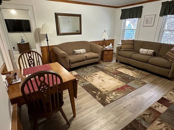 A furnished living room with a wooden dining table, two sofas, a rug, and a television mounted on the wall.