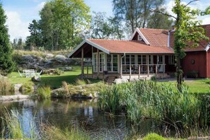 A house with a porch, a pond, and a red building in the background.