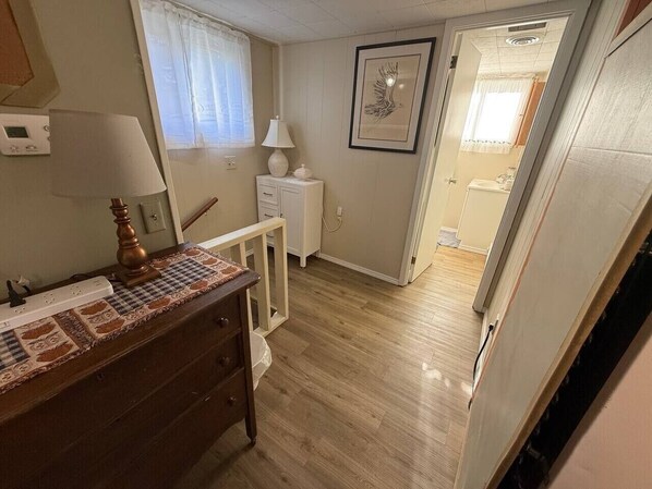 A room with a wooden dresser, a lamp, a framed artwork on the wall, and a door leading to another room.