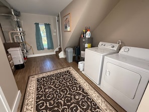 A laundry room with two washing machines, a dryer, a rug, and a window with curtains.