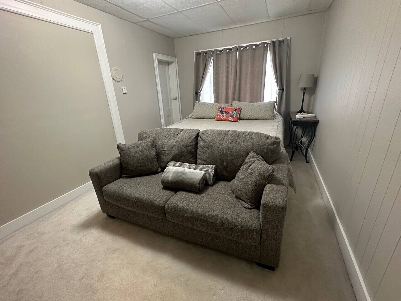 Une chambre avec un canapé gris, un lit à baldaquin, une table de chevet avec une lampe et une porte.