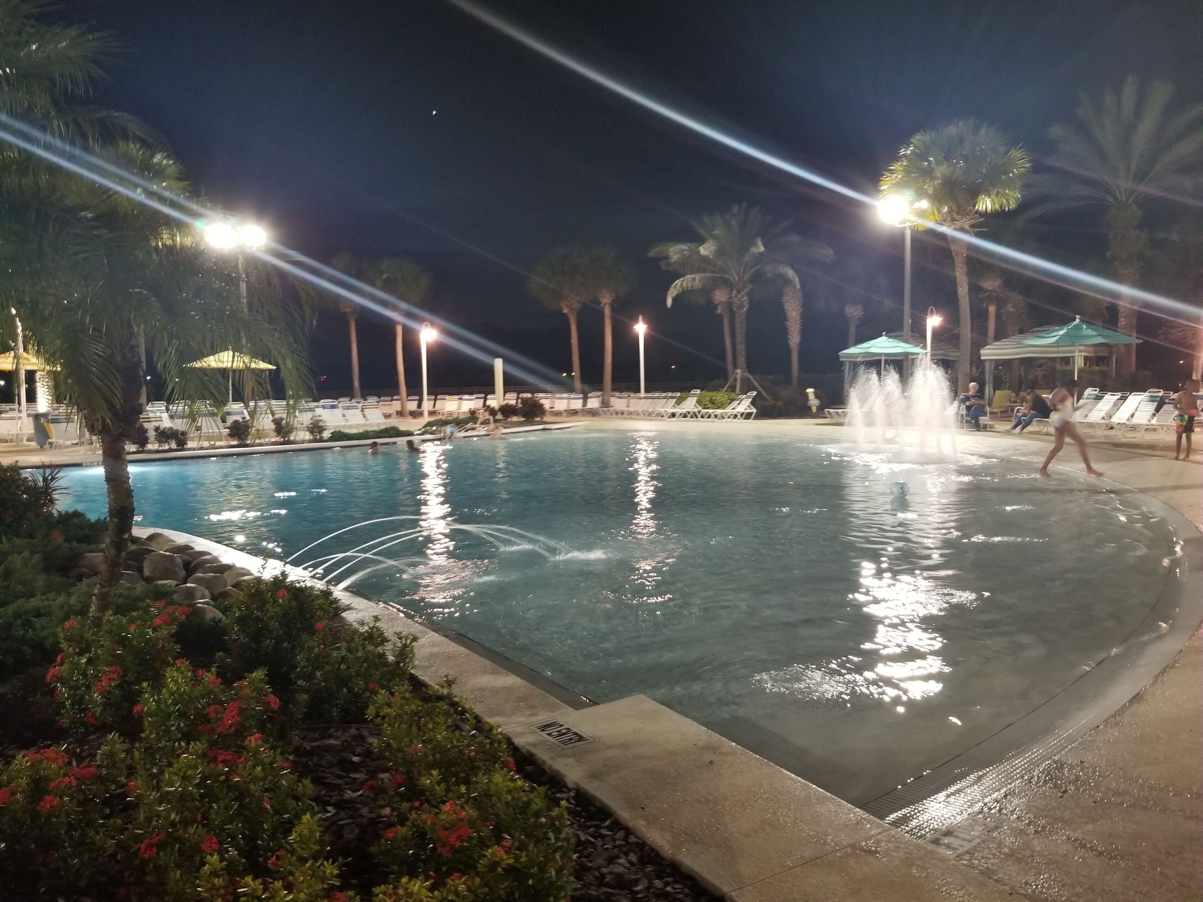 Un espace piscine aménagé pour la nuit, avec une fontaine, des palmiers et des chaises longues.