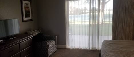 A hotel room with a bed, a television on a dresser, a chair, and a window with sheer curtains.