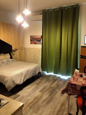 A room with a bed, a table with a checkered tablecloth, a chair, a lamp, and a green curtain. - Vrbo Property (Verona)