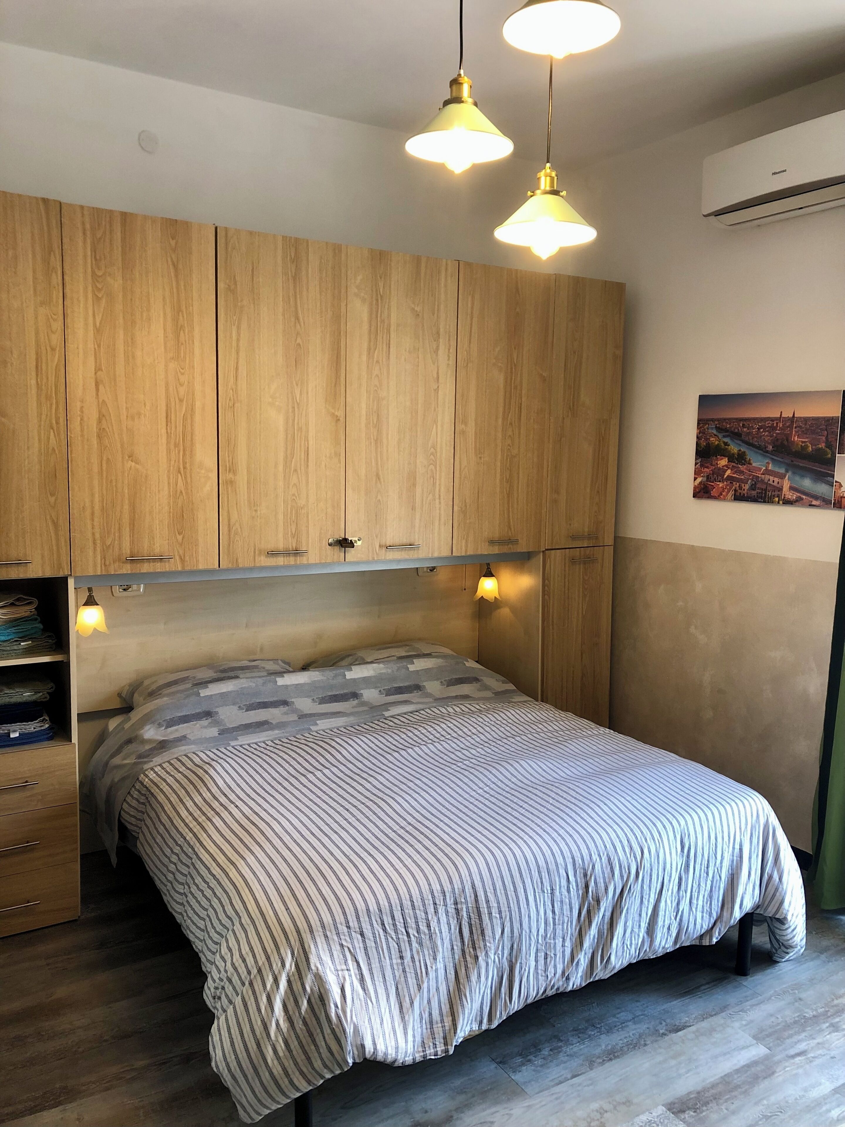 Camera da letto con un letto, un'armadio in legno, luci a sospensione e un quadro raffigurante un panorama urbano.