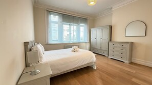 Ein Schlafzimmer mit Bett, Nachttisch, Lampe, Kleiderschrank und einem großen Fenster mit Jalousien.