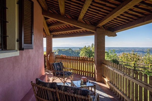 Un espacio de estar al aire libre techado con mobiliario de mimbre y una mesa de vidrio, con vistas a un paisaje pintoresco.