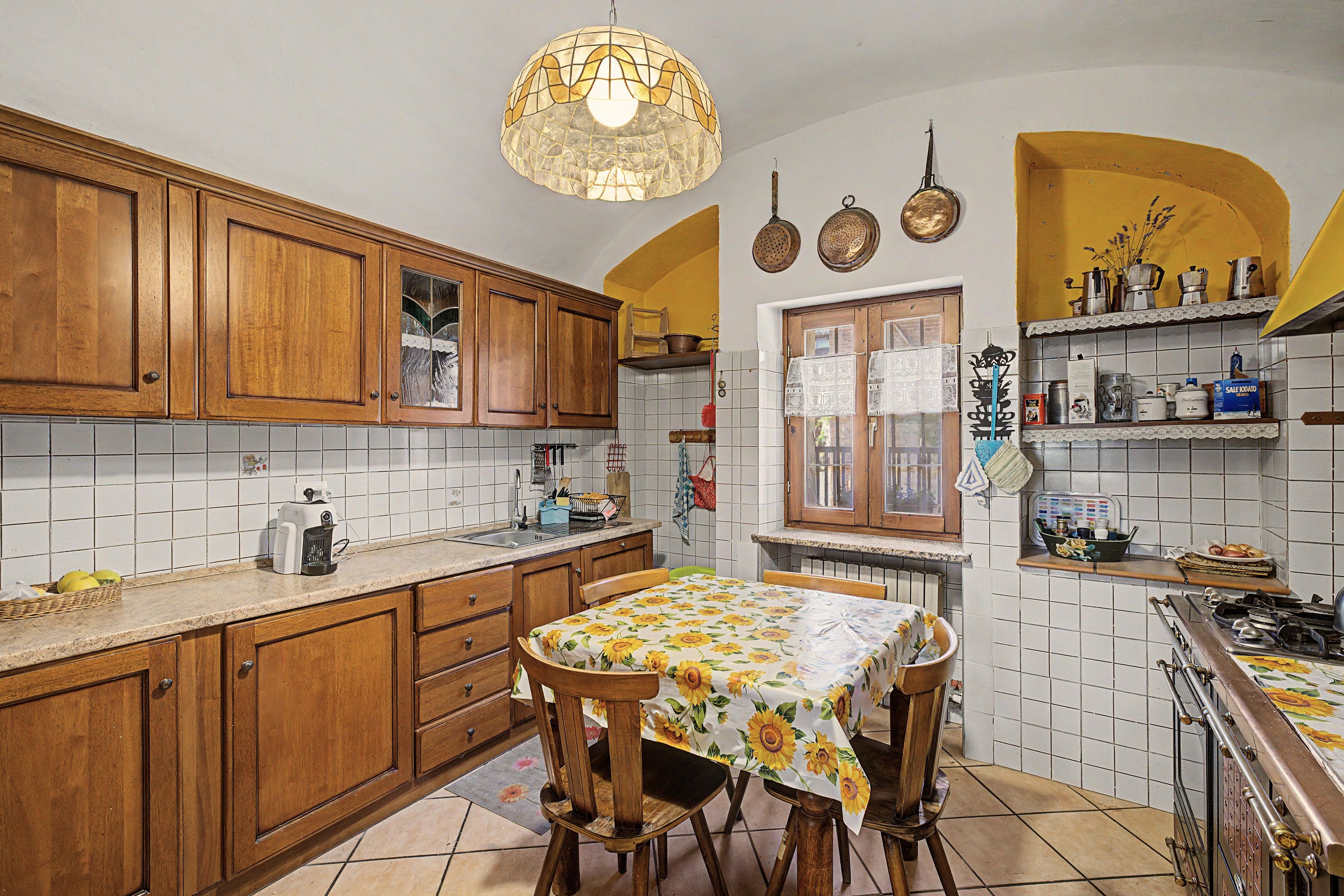 A kitchen with wooden cabinets, a tiled backsplash, a dining table with a floral tablecloth, and a hanging light fixture.