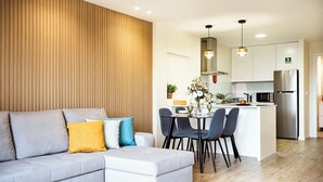 A modern living room with a grey sectional sofa, a dining table with chairs, and a kitchen area with a refrigerator and pendant lights. - Cozy One-Bedroom Retreat with Private Patio & Sunrise Views in Madeira (Ponta do Sol)