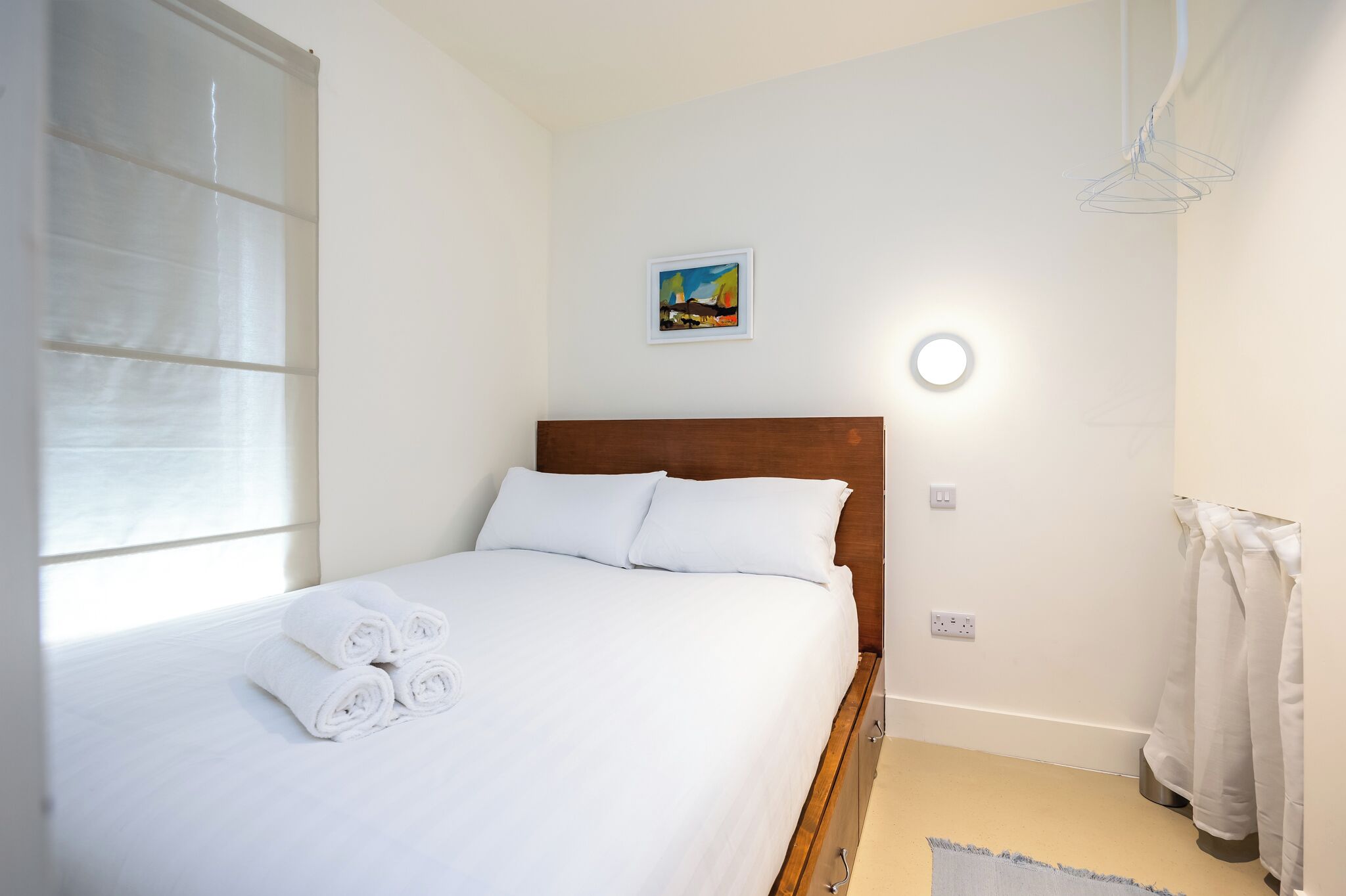 A neatly made bed with white linens and rolled towels, a wooden headboard, a small painting on the wall, and a round wall light.