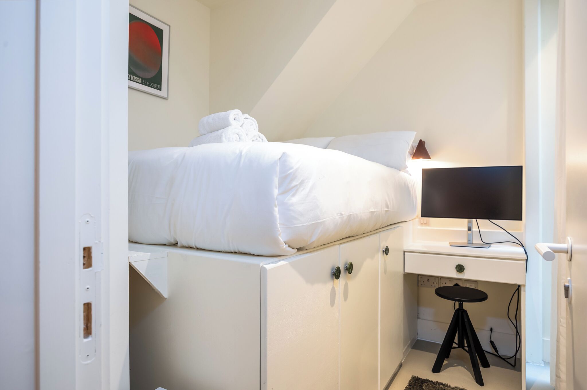 A compact bedroom with a bunk bed, a desk with a computer, and a small stool.