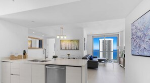 Cozinha moderna com lava-louças embutida, sala de jantar com mesa e cadeiras brancas e sala de estar com sofá cinza e vista para o mar.