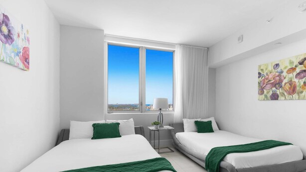 Habitación de hotel con dos camas, una ventana y vista a la ciudad, una mesita con lámpara y obras de arte en las paredes.
