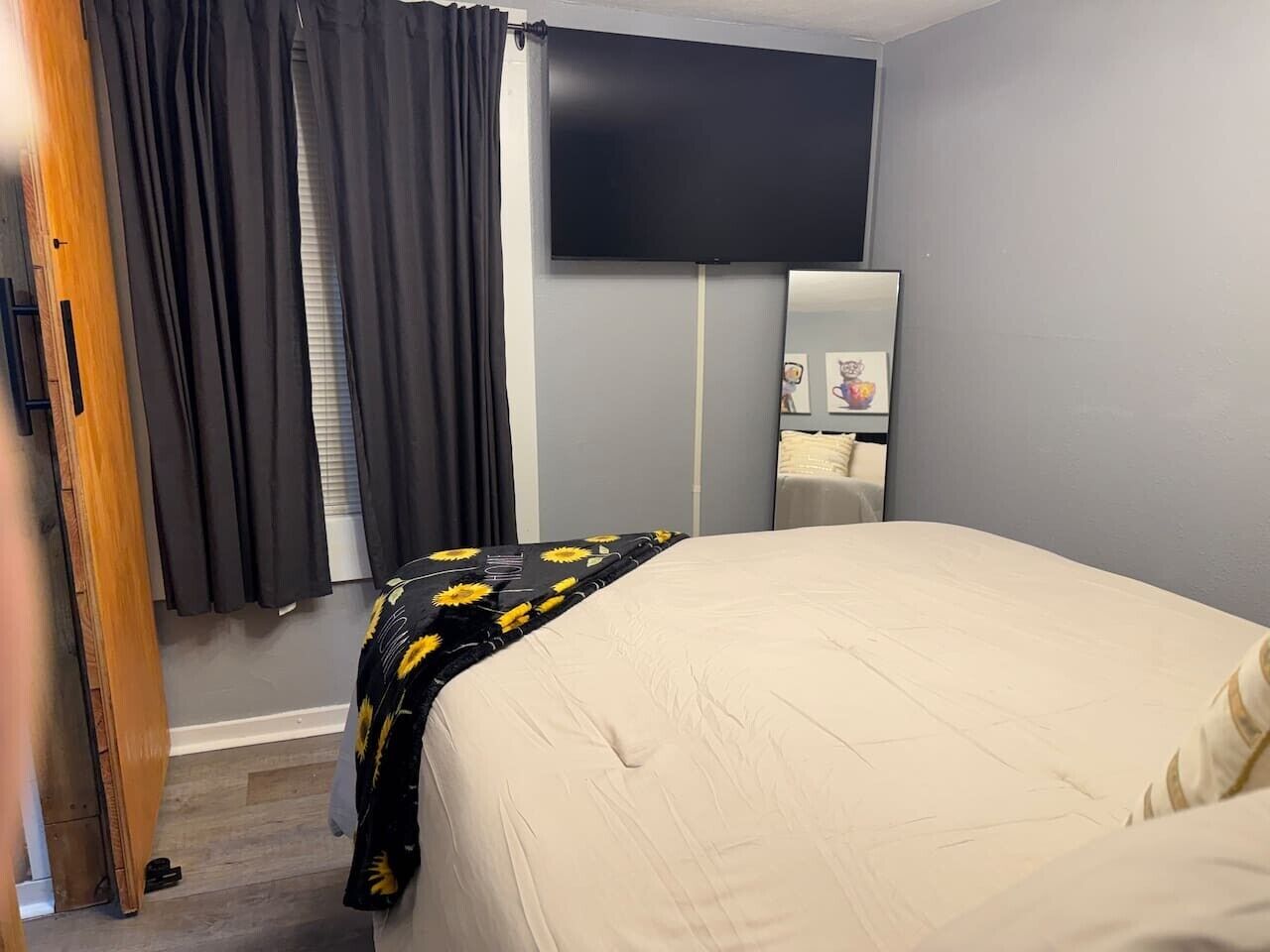 Ein Schlafzimmer mit einem Bett, einem an der Wand befestigten Flachbildfernseher, einem Nachttisch und einem fenster mit Vorhängen.
