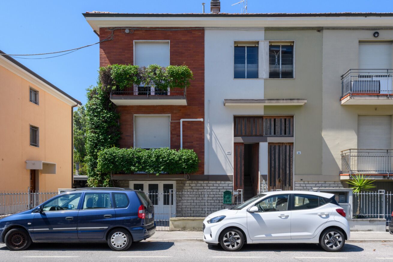 A two-story residential building with a brick facade, a balcony with plants, and a white car parked in front.