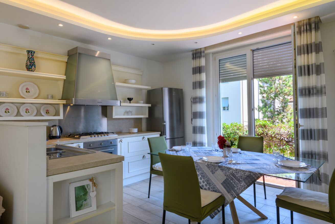 A modern kitchen with a gas stove, oven, and stainless steel appliances. There is a dining area with a table set for two, chairs, and a vase with flowers.