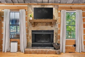 Interior de una cabaña de troncos con chimenea de piedra, un televisor de pantalla plana montado encima y una ventana con cortinas a ambos lados.