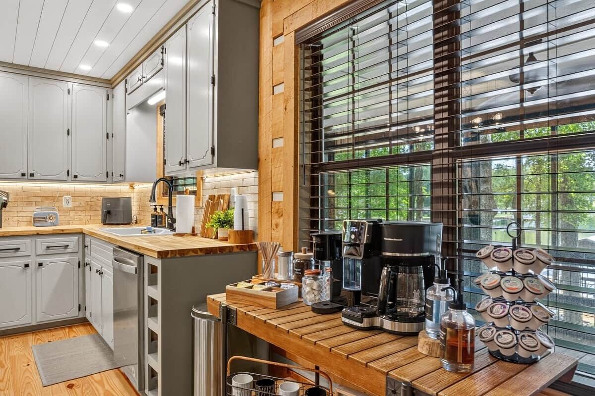A modern kitchen with grey cabinets, a wooden countertop, a coffee station, and a large window with blinds.