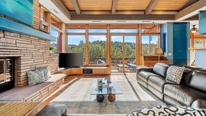 A modern living room with a fireplace, television, leather sofa, and large windows offering a view of the outdoors. - Palisades Villa | 360 Views, Hot Tub, Mid-Century (Sedona)