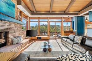 Ein modernes Wohnzimmer mit Kamin, Fernseher, Ledersofa und großen Fenstern mit Blick ins Freie.
