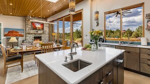 A modern kitchen with a large island, stone fireplace, and large windows offering a view of the outdoors. - Rancho Rojo | Red Rock Views, Hot Tub, Private (Sedona)