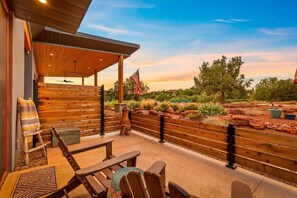An outdoor patio with wooden furniture, a flag, and a view of a landscaped garden. - Rancho Rojo | Red Rock Views, Hot Tub, Private (Sedona)