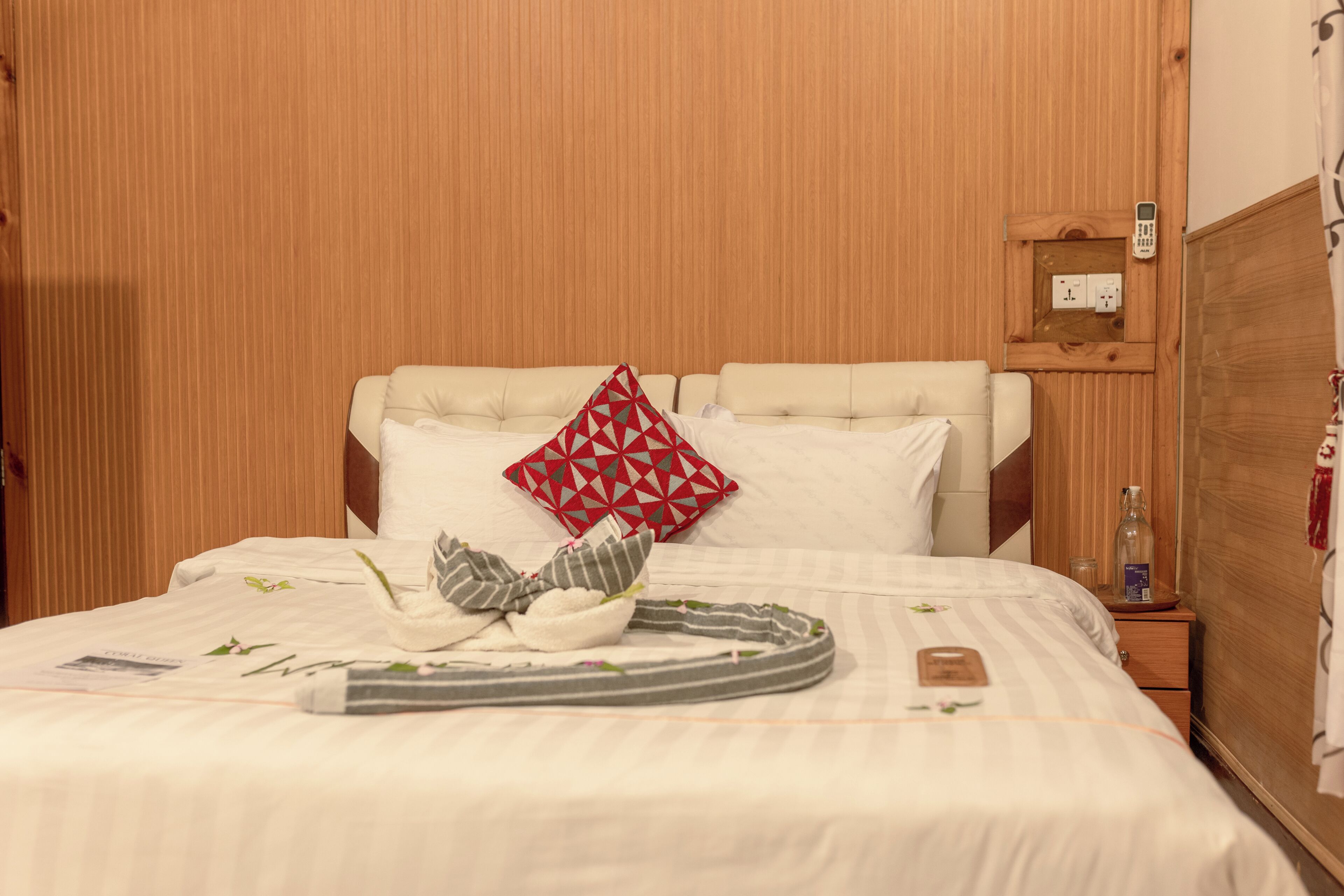 Una cama bien hecha con un cojín a rayas, un cojín geométrico rojo y un cojín blanco. Hay una mesita de noche con una botella y un control remoto.