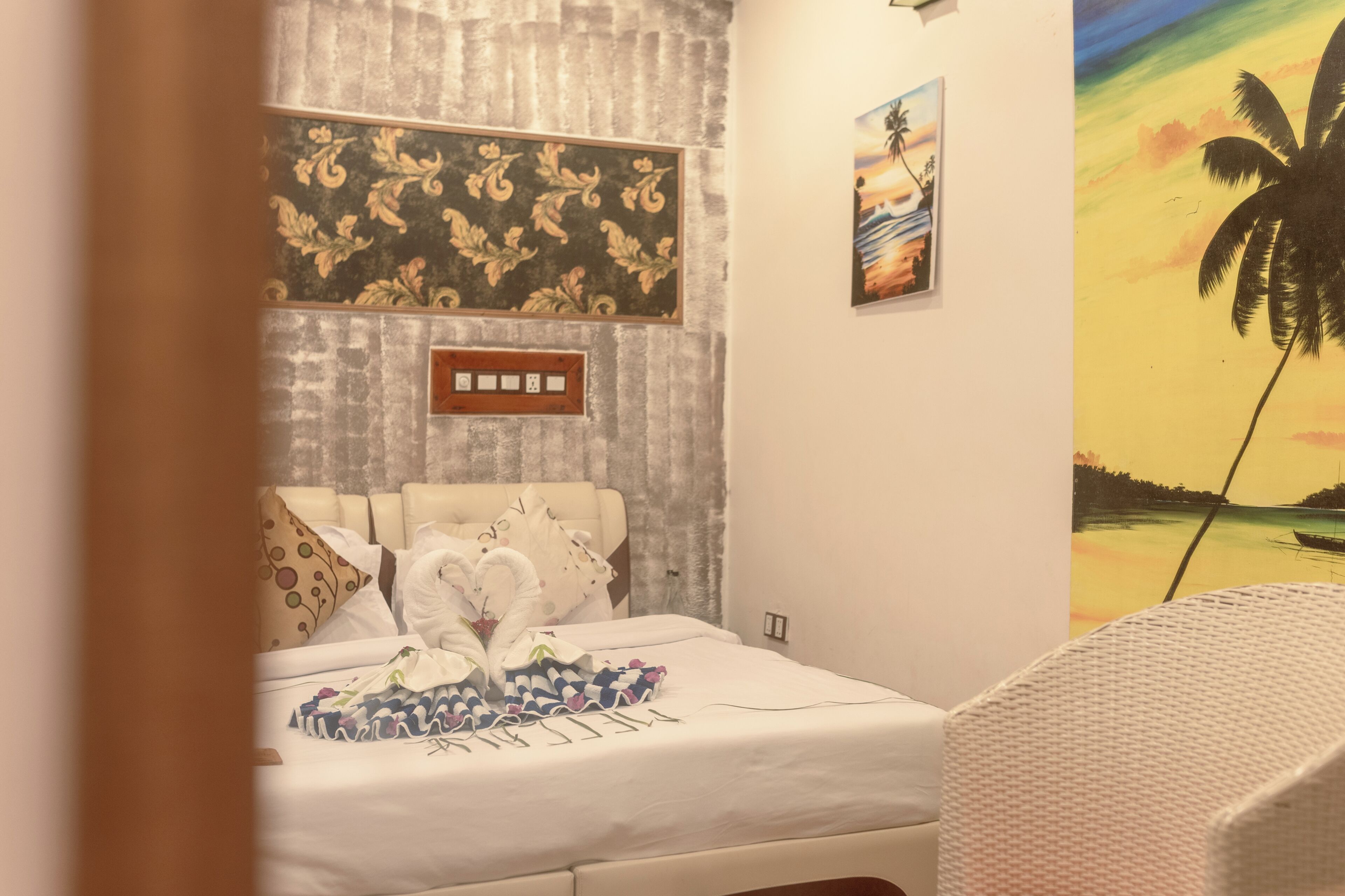 A bedroom with a bed, a wall-mounted power outlet, and a painting of a tropical scene.