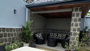 An outdoor patio with a stone wall, a metal roof, and potted plants. - Apartments - Furnished Condos - Yassa Douala - WILMA HOUSE: Just Like Home (Yassa)