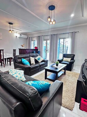 A modern living room with a black leather sofa, a coffee table, and a dining area with a table and chairs. - Apartments - Furnished Condos - Yassa Douala - WILMA HOUSE: Just Like Home (Yassa)