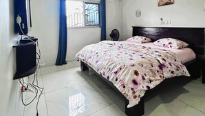 A bedroom with a bed, a TV, a window with curtains, and an air conditioning unit. - Apartments - Furnished Condos - Yassa Douala - WILMA HOUSE: Just Like Home (Yassa)