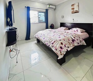 A bedroom with a bed, a TV, a window with curtains, and an air conditioning unit. - Apartments - Furnished Condos - Yassa Douala - WILMA HOUSE: Just Like Home (Yassa)