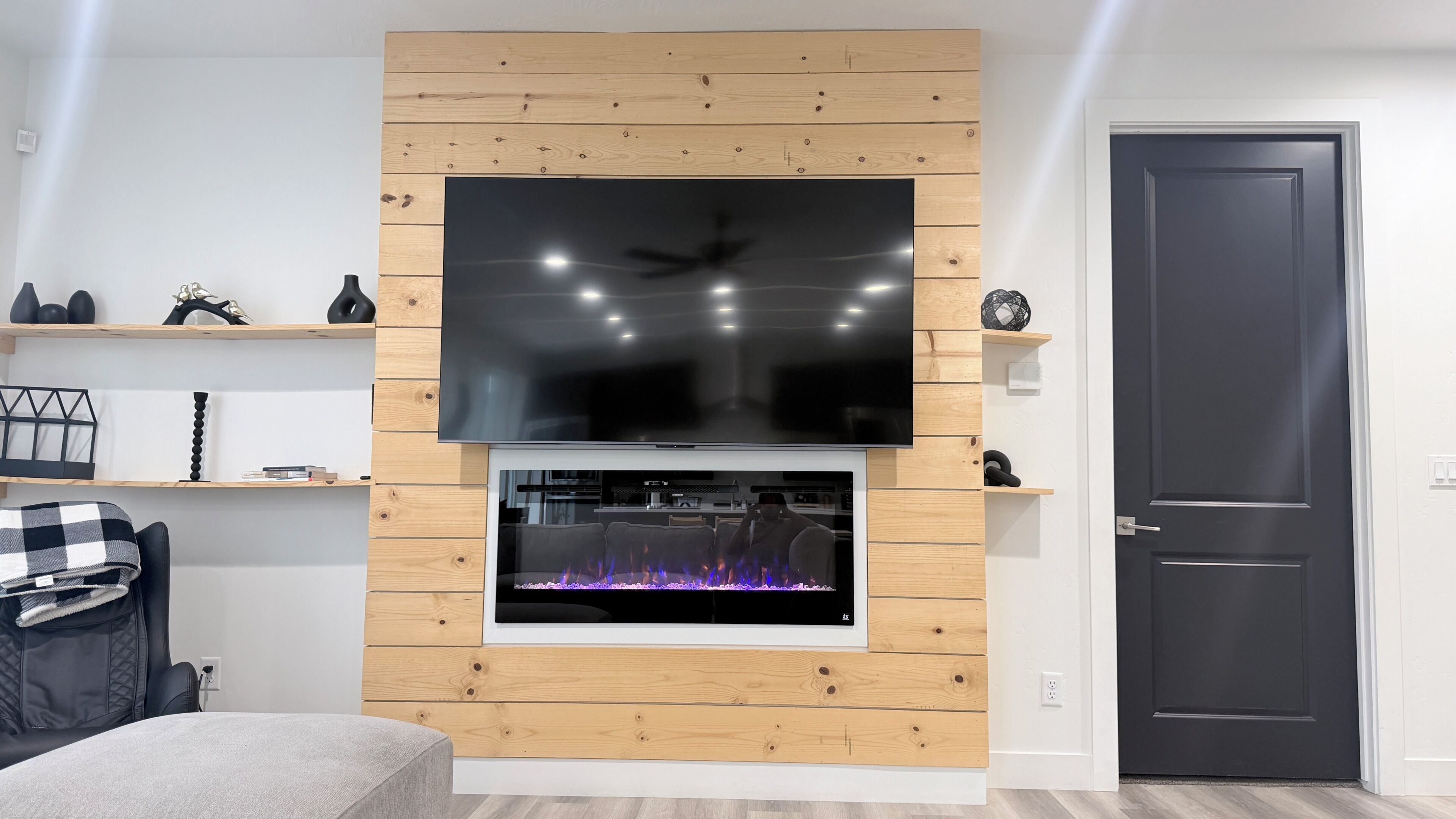 Ein modernes Wohnzimmer mit einem wandmontierten Flachbildfernseher, einem Kamin darunter und einer schwarzen Tür rechts.