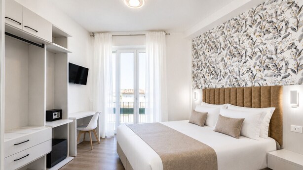 Habitación de hotel con cama, escritorio, televisor y balcón con cortinas.
