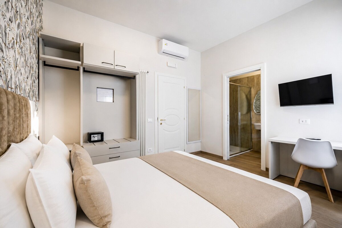 Une chambre d’hôtel comprenant un lit, un bureau, une chaise, une armoire, un téléviseur et une porte donnant accès à une cabine de douche.