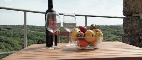 Una mesa de madera con una botella de vino, dos copas de vino y un cuenco de fruta en un balcón con vistas a un viñedo.