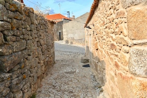 Uma rua de paralelepípedos estreita, ladeada por muros de pedra, que conduz a um edifício com telhado de telhas vermelhas.