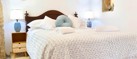 A neatly made bed with a patterned bedspread, two bedside lamps, a bedside table with a woven basket, and a potted plant on the wall.
