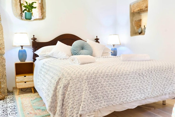 A neatly made bed with a patterned bedspread, two bedside lamps, a bedside table with a woven basket, and a potted plant on the wall.