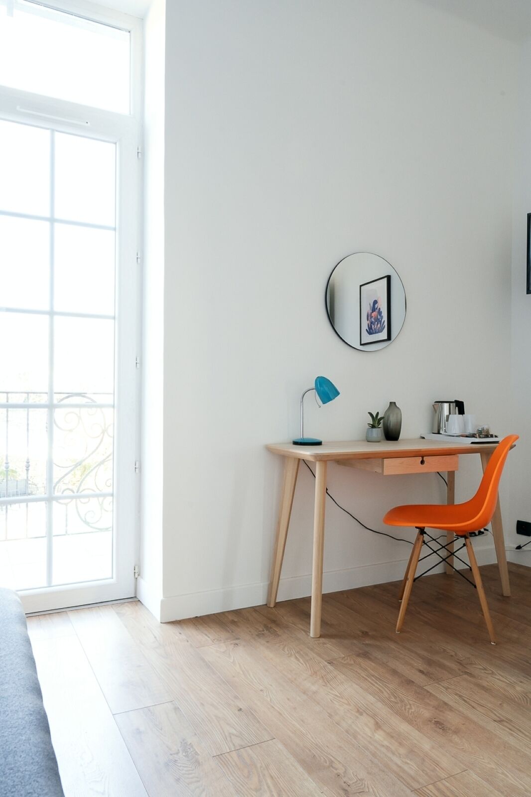 Une pièce avec un bureau en bois, une lampe de bureau bleue, une chaise orange, une petite plante en pot et un miroir rond accroché au mur.