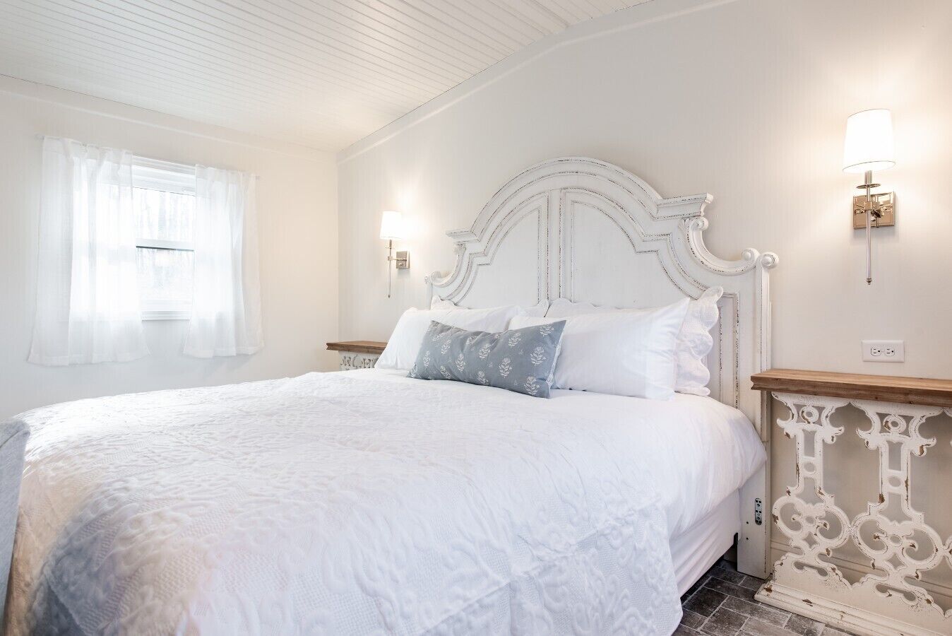 A white bedroom with a large bed, a wooden nightstand, and a wall-mounted lamp.
