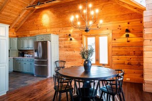 A wooden cabin interior with a dining area, kitchen, and a chandelier. - Secluded 51-Acre Retreat | 2 Cabins, Pond & Garden (Henryville)