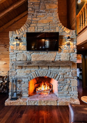A stone fireplace with a TV mounted above it, a fire burning inside, and a wooden ceiling.