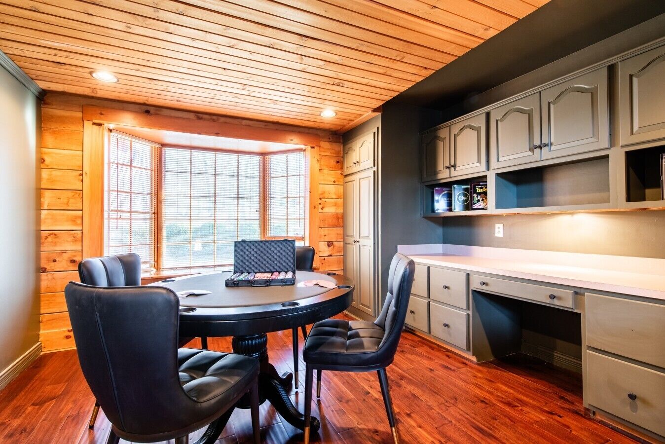 A room with a round table, two chairs, a window with blinds, wooden walls, and a kitchenette with cabinets.